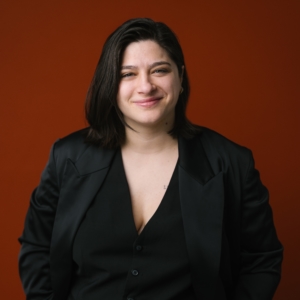 Sorority Board Member Sibel Ada, Porträtfoto in schwarzem Blazer vor orangefarbenen Hintergrund